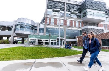Two UVU students walk by Fulton Library, with a library entrance and two other students in the background
