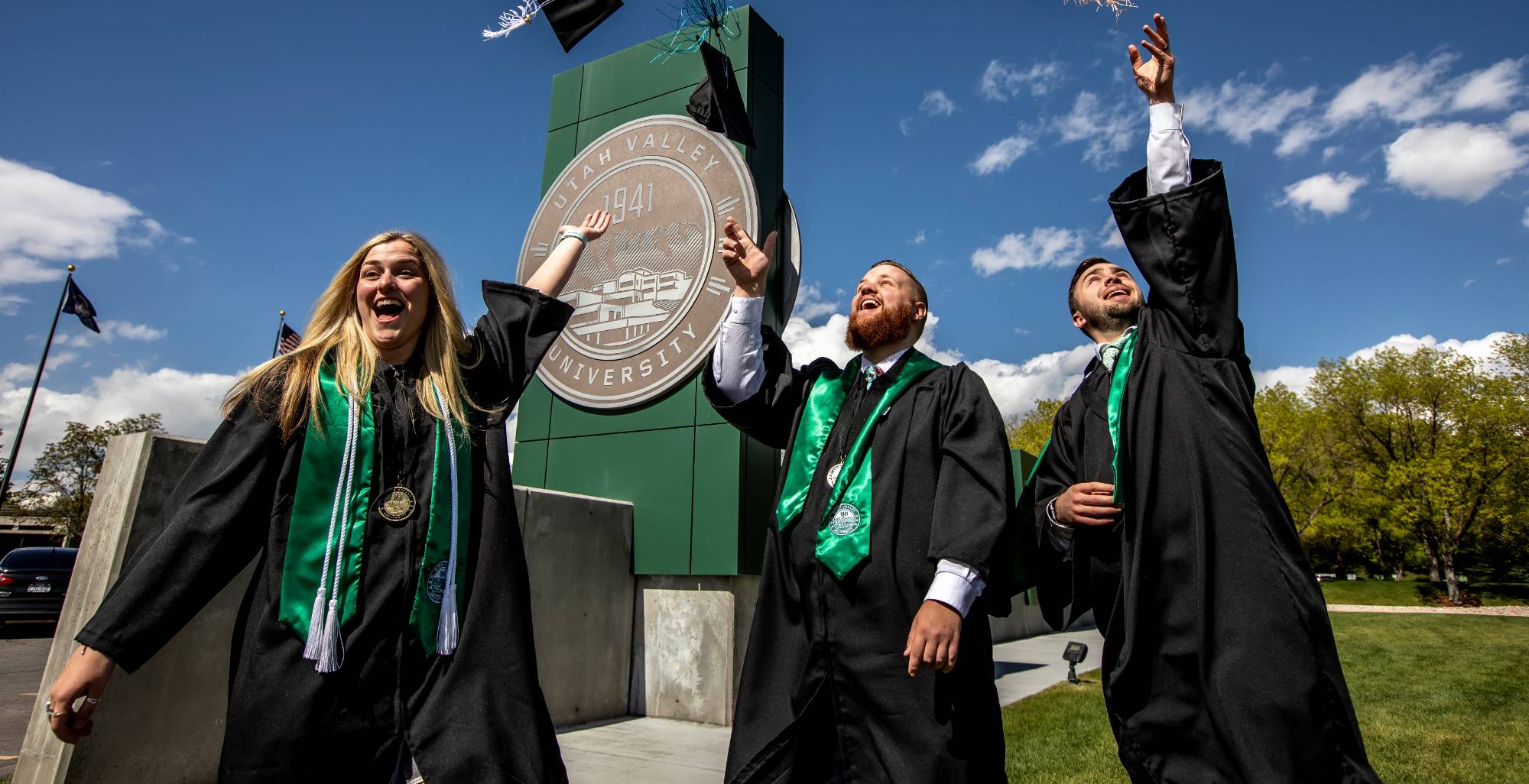 3 students throwing caps up at graduation in front of UVU sign