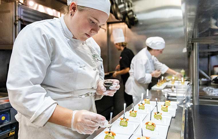 A UVU Culinary Arts student working in a kitchen adds finishing touches to hors d'oeuvres on white plates. 