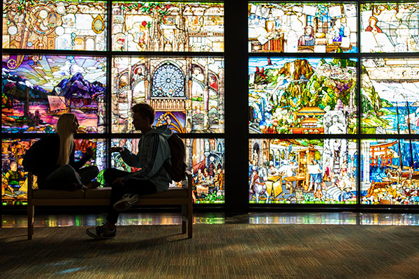 2 people at the UVU Roots of Knowledge Room, where there is stain glass windows in the background