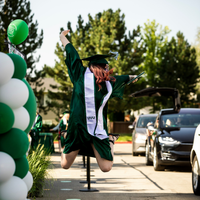 female student in green cap and gown, jumping in the air