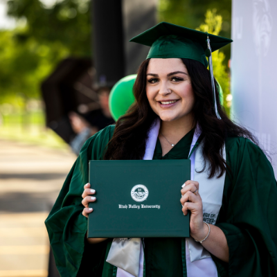 smiling, dark haired female student, in green cap and gown, holding diploma cover