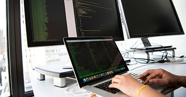 hands on a laptop keyboard, monitors in the background