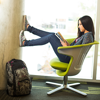 Dark haired female student sitting in a chair with her legs propped up on the wall reading a book