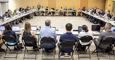 Photo of a Faculty Senate meeting. A large room with tables set in a square with the senate facing inwards towards eachother.