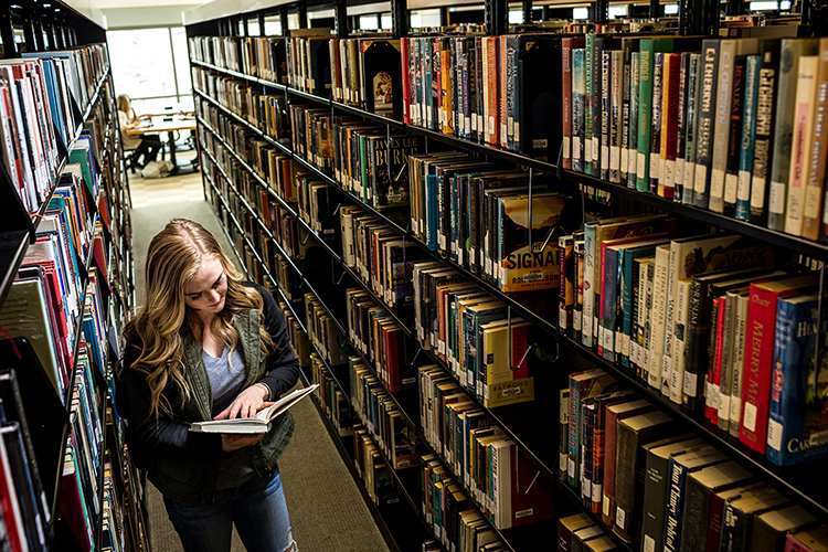 Student in library reading a book between two shelves