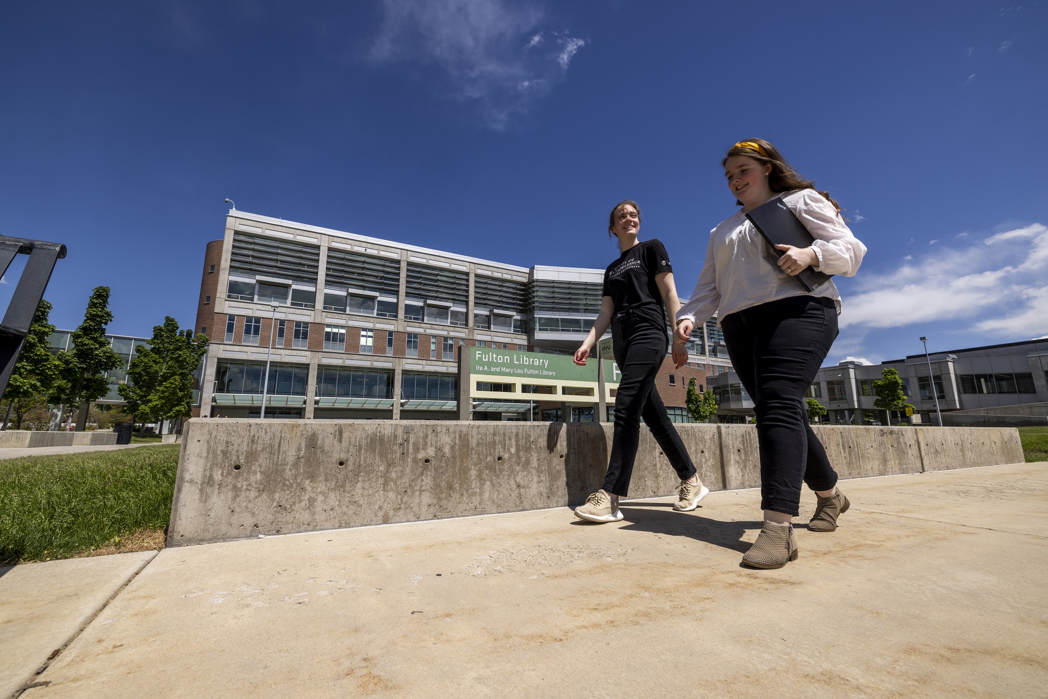 Two people walking on a pathway in front of the modern Fulton Library under a clear blue sky.