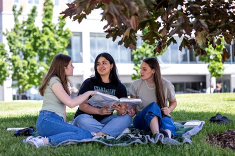 Three women sitting on a blanket on grass, talking and looking at a book.