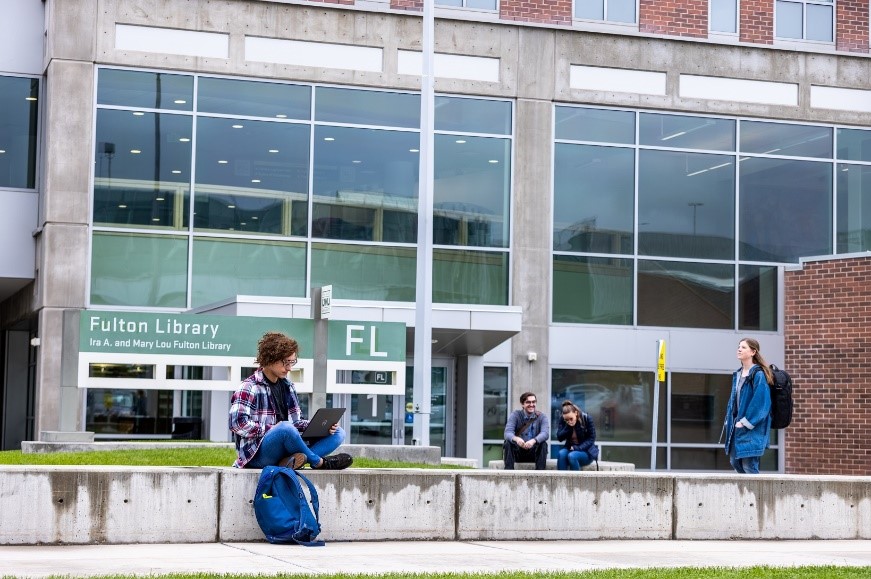 People outside the Fulton Library, with one using a laptop and others walking or sitting.