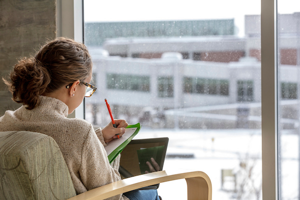 woman next to a window writing in a notebook with laptop on her lap