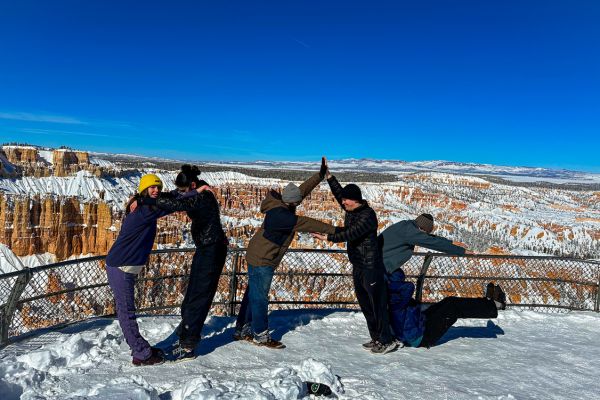 People creating the letters O A C with their bodies with snow-capped red rock formations in the background