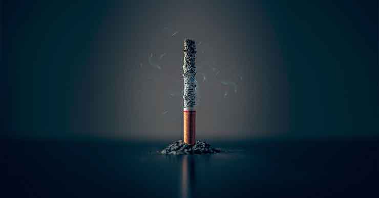 A cigarette standing upright, burning with ash falling on the table.
