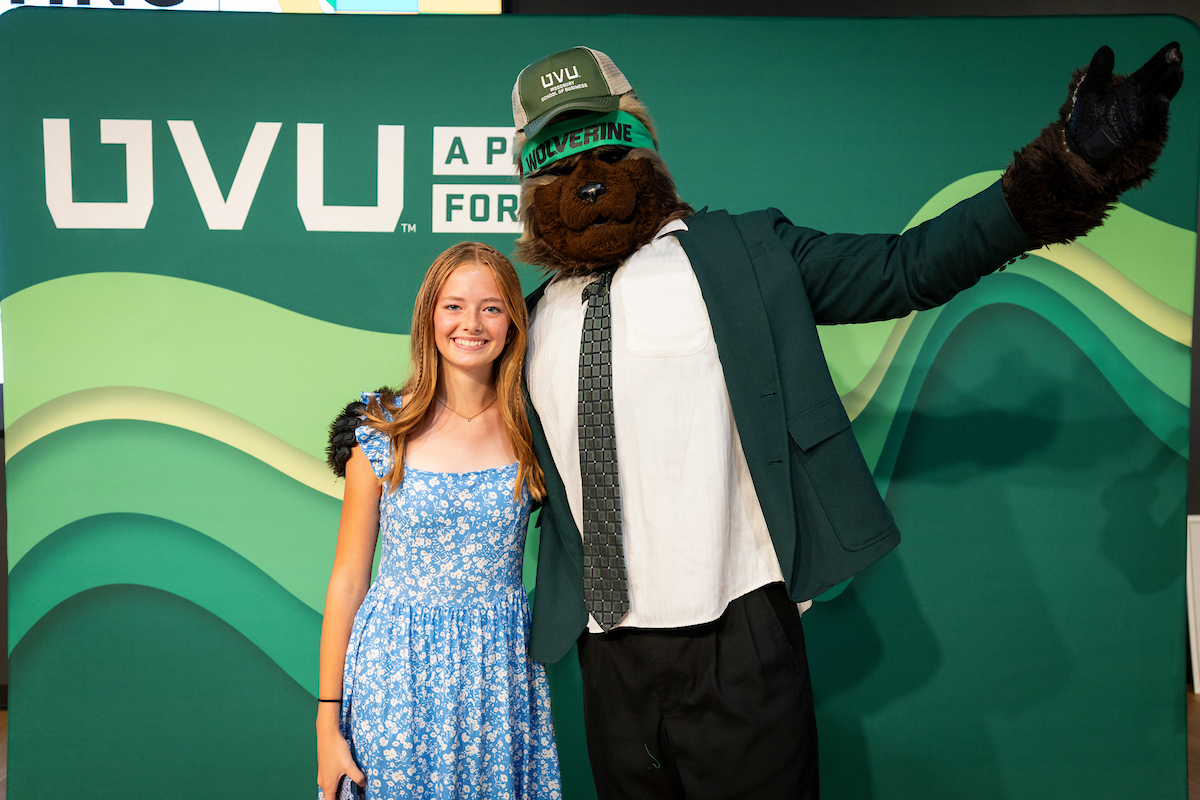 Willy the Wolverine and a female student excited in front of green backdrop with UVU logo